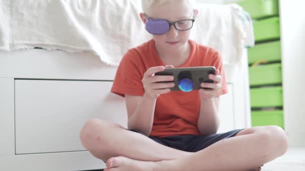Мальчик в очках с приклеенным глазом пользуется мобильным телефоном дома — стоковое видео