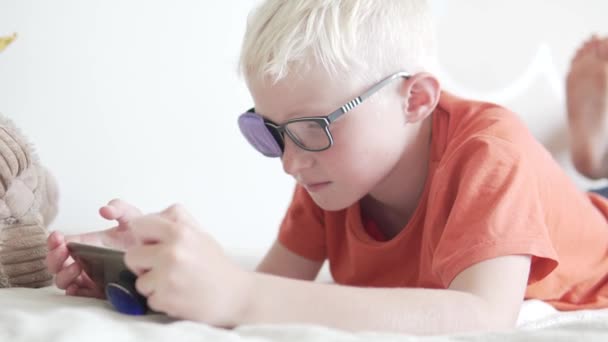 Un niño con gafas con un oclusor mira en el teléfono móvil — Vídeos de Stock