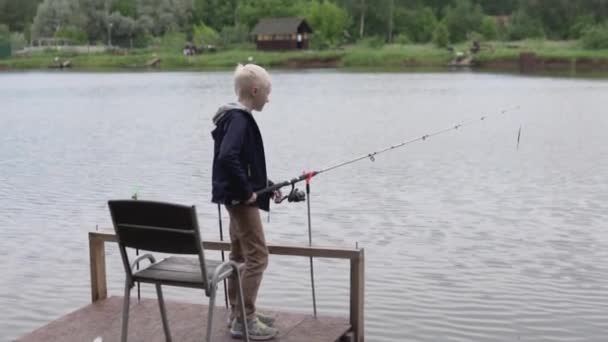 Egy gyönyörű fiú szőke hal horgászbot a mólón a tó mellett nyáron — Stock videók