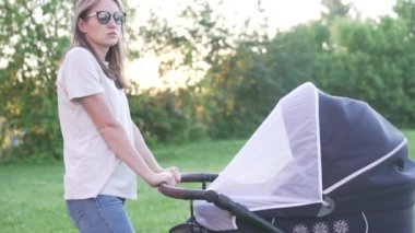 Genç bir anne, yazın gün batımında parkta yeni doğmuş bir bebekle bebek arabasını sallar.