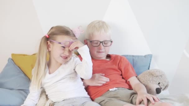 Bruder und seine kleine Schwester mit Brille sitzen auf dem Bett. — Stockvideo