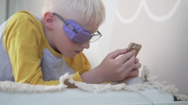 En närbild av en blond pojke med ockluderglasögon spelar i en mobiltelefon — Stockvideo