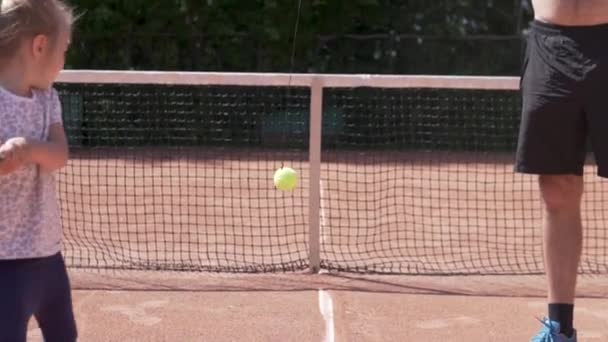 Ένα κοριτσάκι μαθαίνει να παίζει τένις σε ένα βρώμικο γήπεδο το καλοκαίρι. — Αρχείο Βίντεο