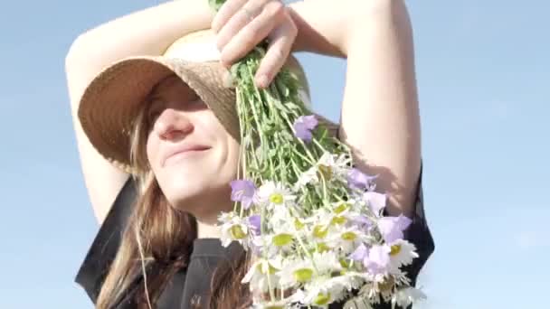 Primer plano de una hermosa mujer en un sombrero de paja con un ramo de flores silvestres en su mano contra el cielo — Vídeos de Stock