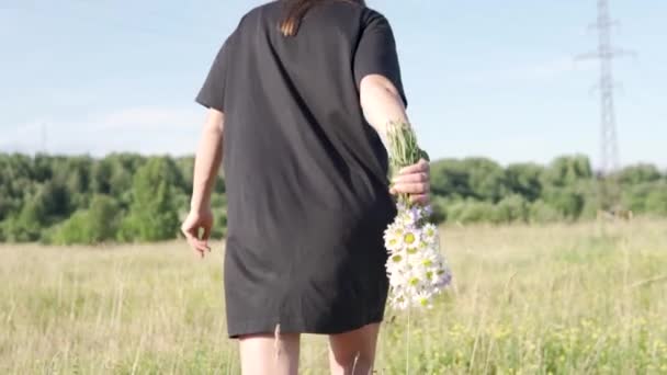 Uma menina com um buquê de flores silvestres atravessa o campo em um vestido preto no verão — Vídeo de Stock