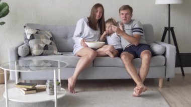 Küçük çocuklu bir aile televizyon izler ve patlamış mısır yer..