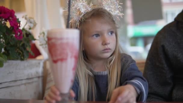 Una niña divertida se sienta en un café con un gran vaso de cóctel de leche de fresa — Vídeos de Stock