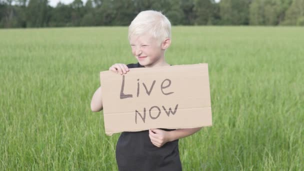 Хлопець білявка стоїть у полі з плакатом на картоні з написом "Live NOW — стокове відео