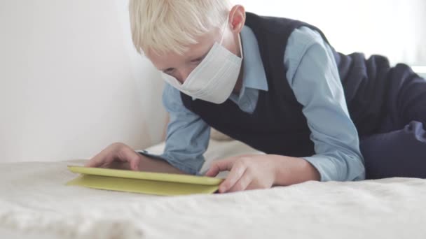 Um menino de escola com uma máscara médica em seu rosto joga em um tablet em casa — Vídeo de Stock