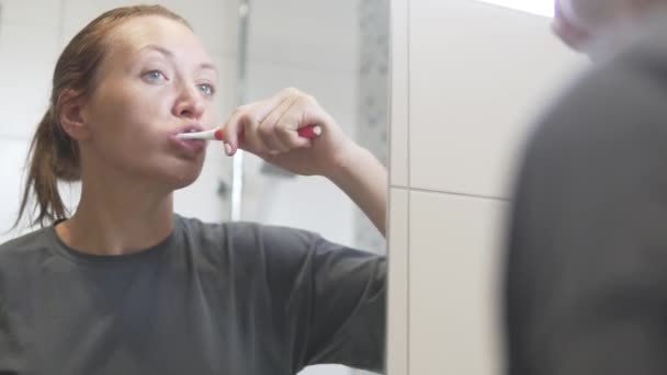 Bir kadın sabah dişlerini banyoda aynanın önünde fırçalar. — Stok video