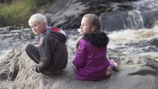 Chłopiec i nastolatka siedzą na kamieniu i patrzą na wodospad. — Wideo stockowe