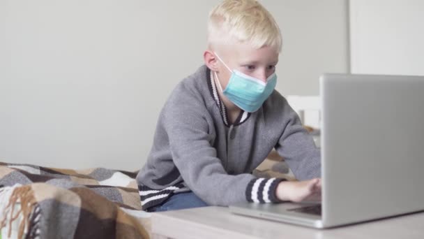 Een jongen met een medisch masker is thuis met een computer bezig. — Stockvideo