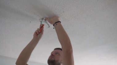 A man connects electrical wires on the ceiling while preparing to install a chandelier. He uses a screwdriver during a home renovation, showing precise indoor electrical work. Ideal concept for DIY