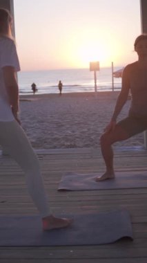 Two people are practicing yoga on mats by the beach during sunrise.