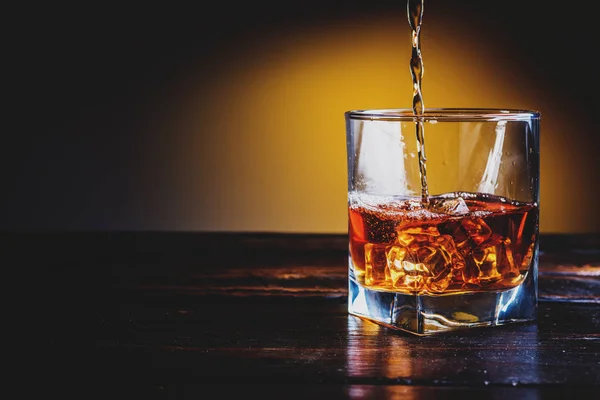 Alcohol drink whisky, whiskey or bourbon with ice cubes on dark wood table and orange background