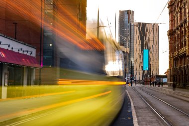 Light rail yellow tram in the city center of Manchester, UK in the evening. Motion blurred tram with modern buildings clipart