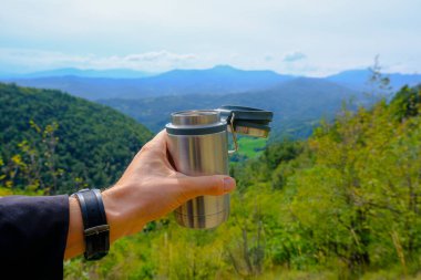 Metal termo bardakta sıcak içecek, insanın ellerinde, dağların manzarasına yakın çekim. Açık hava aktivitesi. Dağlarda yürüyüş / yürüyüş yapmak. Boşluğu kopyala