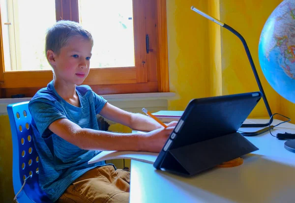 Cute blonde boy with pen and copybook studying using on laptop/tablet at the desk. Educating online/remotely/ homeschooling. Pandemic education.