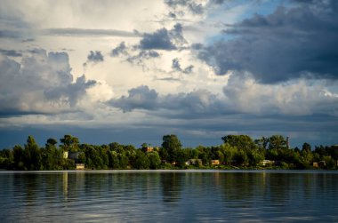 Geniş bir nehri olan bir manzara, binalarla dolu bir orman kıyısı ve beyaz bulutlu yüksek bir gökyüzü..