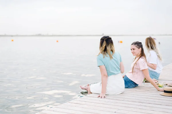 Τρία κορίτσια που κάθονται σε μια ξύλινη προβλήτα μπροστά στο νερό. ανάπαυση, πικνίκ, λεμονάδα — Φωτογραφία Αρχείου