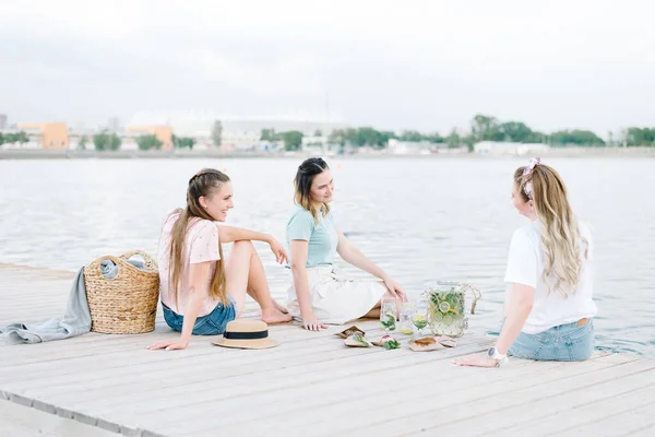 Τρία κορίτσια που κάθονται σε μια ξύλινη προβλήτα μπροστά στο νερό. ανάπαυση, πικνίκ, λεμονάδα — Φωτογραφία Αρχείου