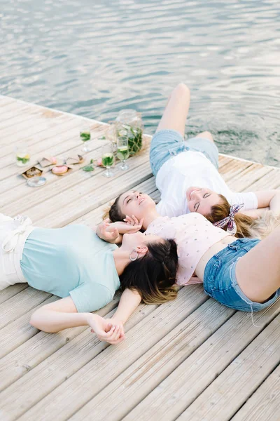 Three girls lie on a wooden pier in front of the water. rest, picnic, lemonade — Stock Photo, Image