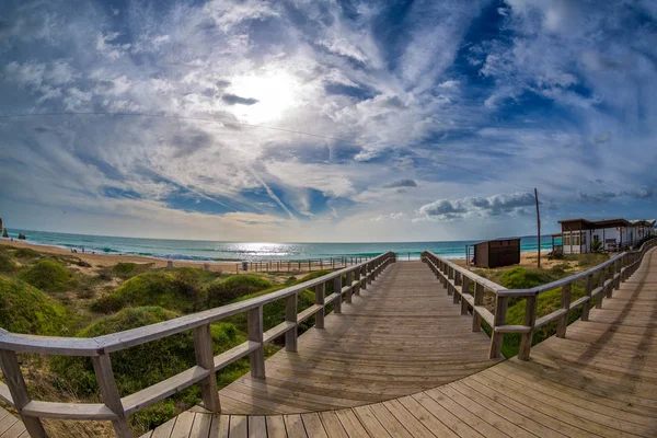 Ahşap yürüyüş yolları Alvor, Portimao, Algarve, Portekiz, Avrupa ünlü Tres Irmaos Beach'te okyanusta yol. Praia dos Tres Irmaos. Kuma inişli çıkışlı dalgalar bulutların arasından güneş parlıyor.