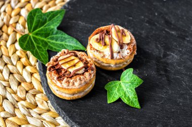 İki pasta bisküvi ile beyaz kremalı muz dilimleri ve çikolata, mazisi bir hasır masa mat arka plan üzerinde çanağı. Yeşil sarmaşık yaprakları ile süslenmiş. Üstten görünüm. Metin için boş alan.