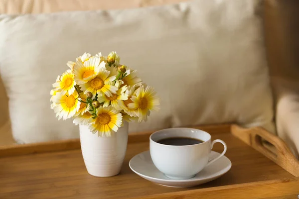 Bir fincan çay / kahve ve bir buket bir ahşap tepsi hafif yumuşak yastık arka planı üzerinde bir vazoda papatyalar