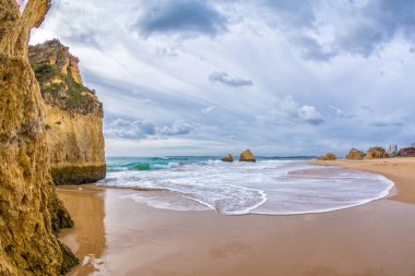 Ünlü kaya oluşumu bir koyda Alvor, Portimo, Algarve, Portekiz, Avrupa'nın Tres Irmaos plaj üzerinde. Praia dos, Tres Irmaos.The Güneş bulutların arasından parlıyor dalgalar kumsalda rulo. İki kayaların arasına görüntülemek