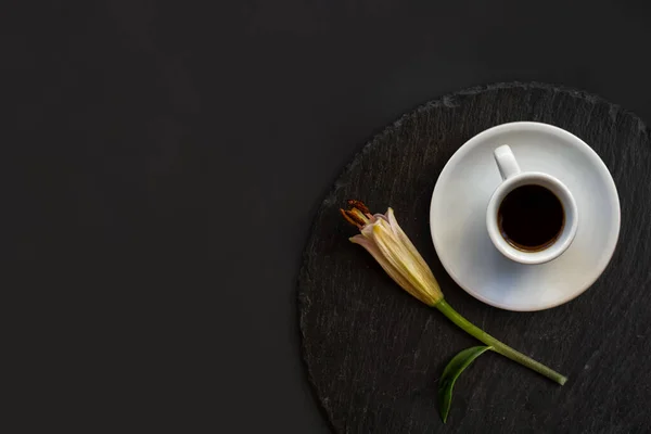 Cup of black coffee and fresh spring single bud of lily flower on a round slate on a black background. Romantic stylish elegance breakfast. Date on restaurant. Still life flat lay. Top view. Space for text.