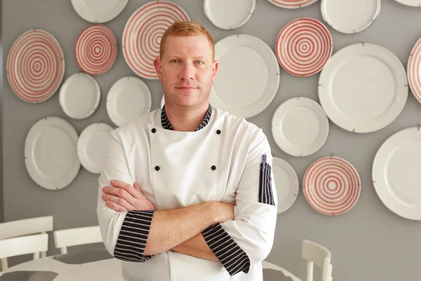 Genç, uzun boylu, kızıl saçlı, mavi gözlü aşçı. Kolları çapraz duran aşçı. Restoranının tabakla süslenmiş duvarının arkasında duruyor.