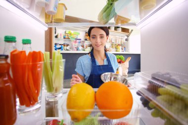 Sağlıklı yiyecek, sebze ve meyvelerle dolu açık buzdolabının yanında duran kadın portresi. Kadın portresi