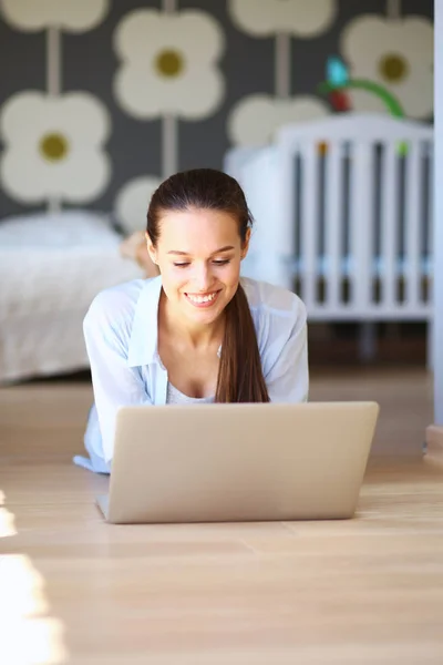 Ung kvinna som sitter på golvet nära barnens babysäng med laptop. Ung mamma — Stockfoto