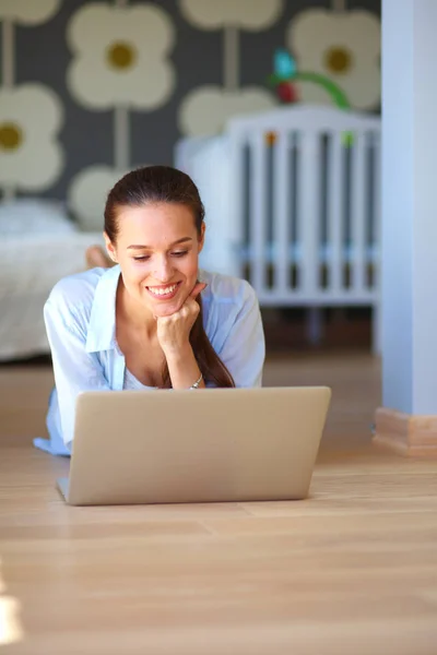 Ung kvinna som sitter på golvet nära barnens babysäng med laptop. Ung mamma — Stockfoto