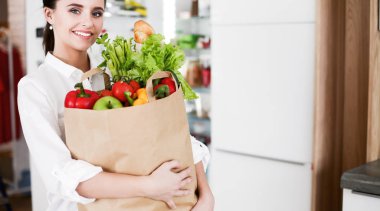 Sebzeli alışveriş torbası tutan genç bir kadın. Mutfakta duruyor. Mutfakta kameraya bakan bir kadın var.