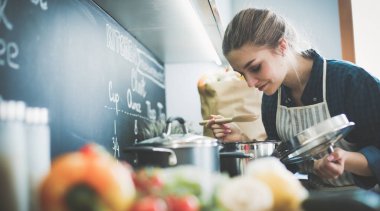 Genç bir kadın mutfağında yemek pişiriyor sobanın yanında duruyor.