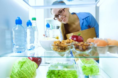 Sağlıklı yiyecek, sebze ve meyvelerle dolu açık buzdolabının yanında duran kadın portresi. Kadın portresi