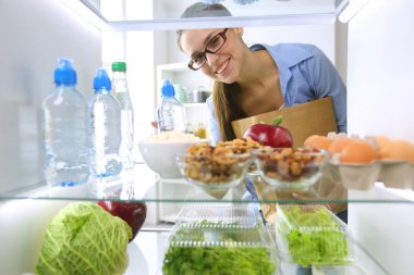 Sağlıklı yiyecek, sebze ve meyvelerle dolu açık buzdolabının yanında duran kadın portresi. Kadın portresi
