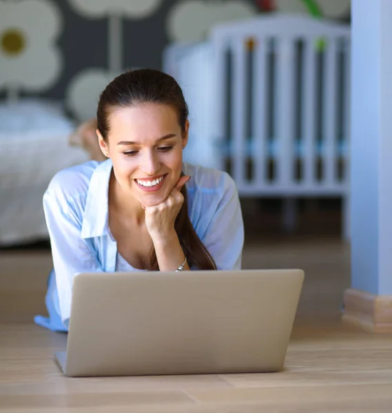 Ung kvinna som sitter på golvet nära barnens babysäng med laptop. Ung mamma — Stockfoto