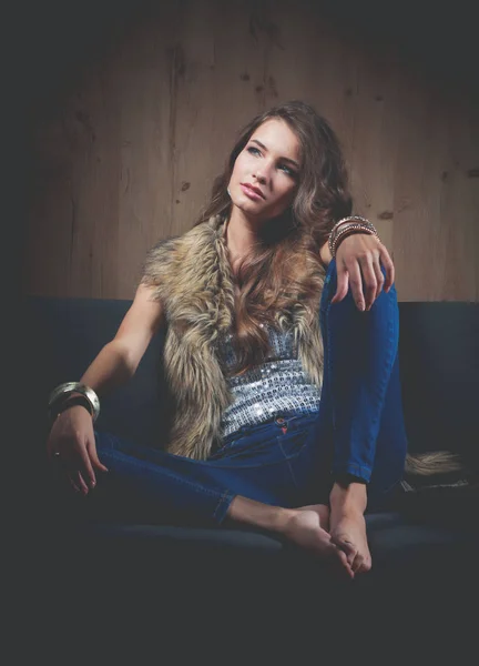 Portrait of elegant woman sitting on black sofa wearing a blue jeans and fur vest — Stock Photo, Image