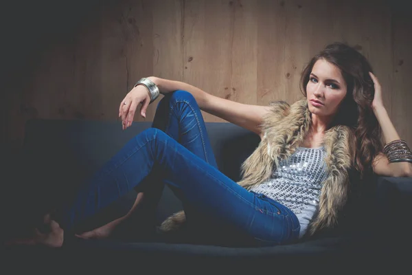 优雅的女人坐在黑色的沙发上，穿着一件蓝色牛仔裤的肖像和毛皮背心 — 图库照片