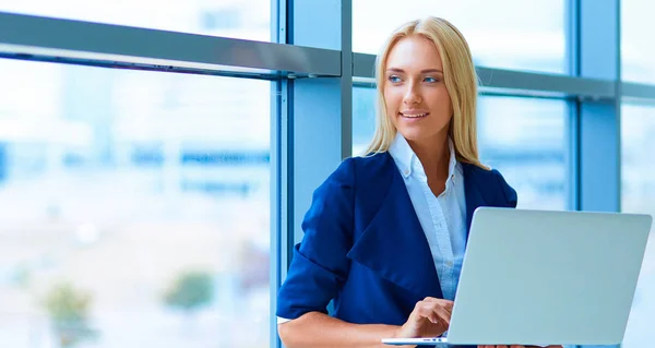 Donna d'affari in piedi contro la finestra dell'ufficio che tiene il computer portatile — Foto Stock