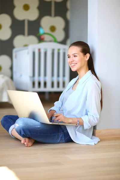 Ung kvinna som sitter på golvet nära barnens babysäng med laptop. Ung mamma — Stockfoto