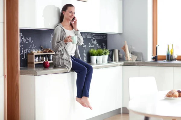 Mujer usando el teléfono móvil sentado en la cocina moderna . — Foto de Stock