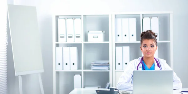 Hermosa joven sonriente doctora sentada en el escritorio y escribiendo. — Foto de Stock