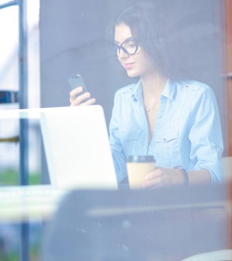 Ofis masasında dizüstü bilgisayarıyla oturan genç bir kadın. Pencereden bakıyor. Genç kadın.