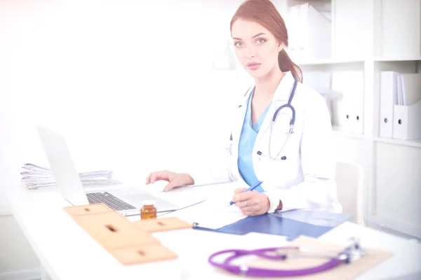 Hermosa joven sonriente doctora sentada en el escritorio y escribiendo — Foto de Stock