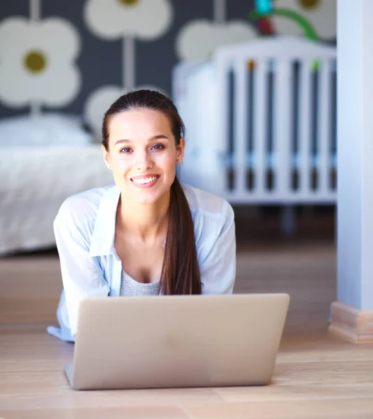 Ung kvinna som sitter på golvet nära barnens babysäng med laptop. Ung mamma — Stockfoto