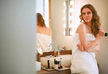 Genç bir kadın aynaya bakıyor ve makyaj yapıyor..